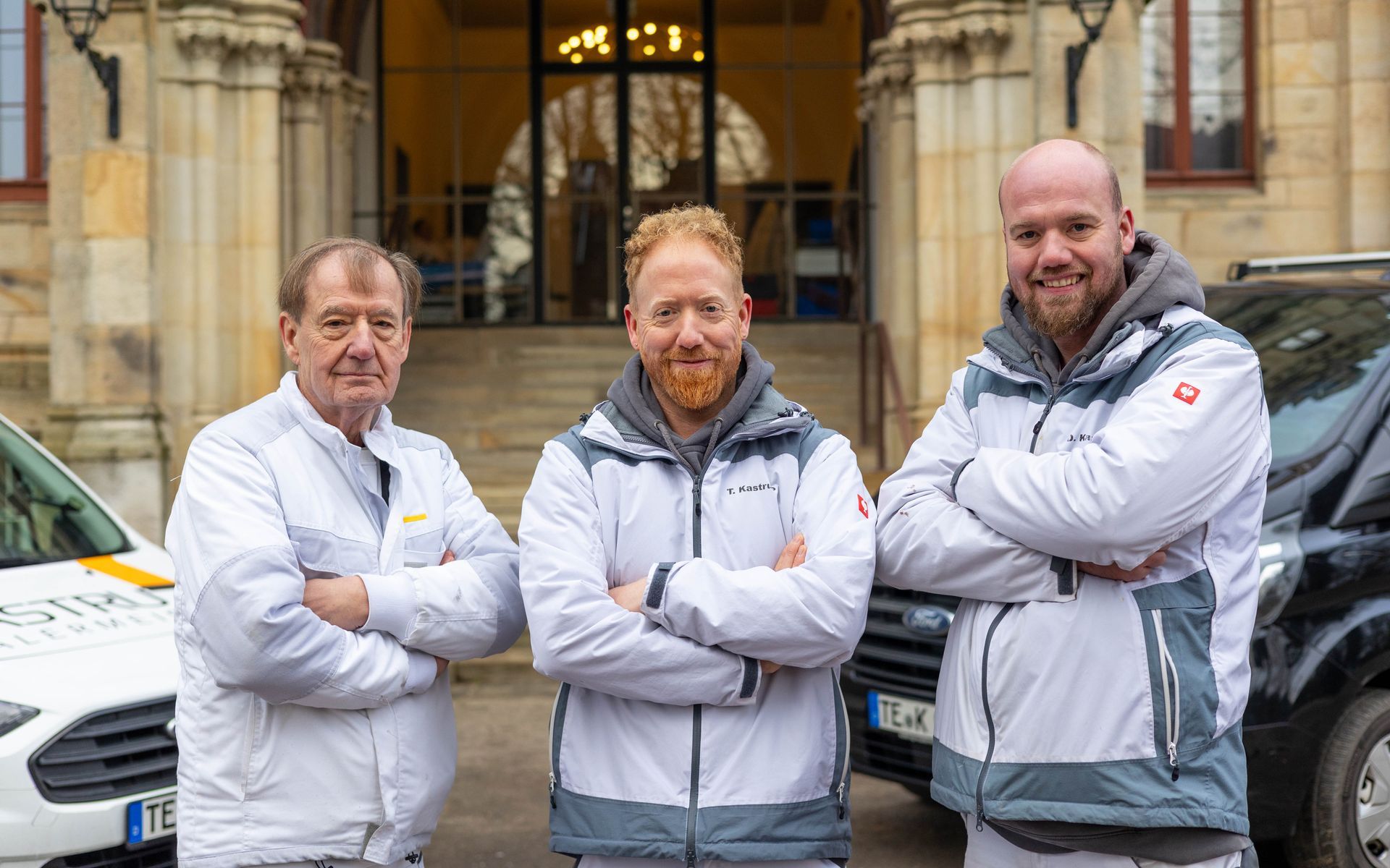 Drei Männer stehen mit verschränkten Armen nebeneinander vor einem Gebäude.