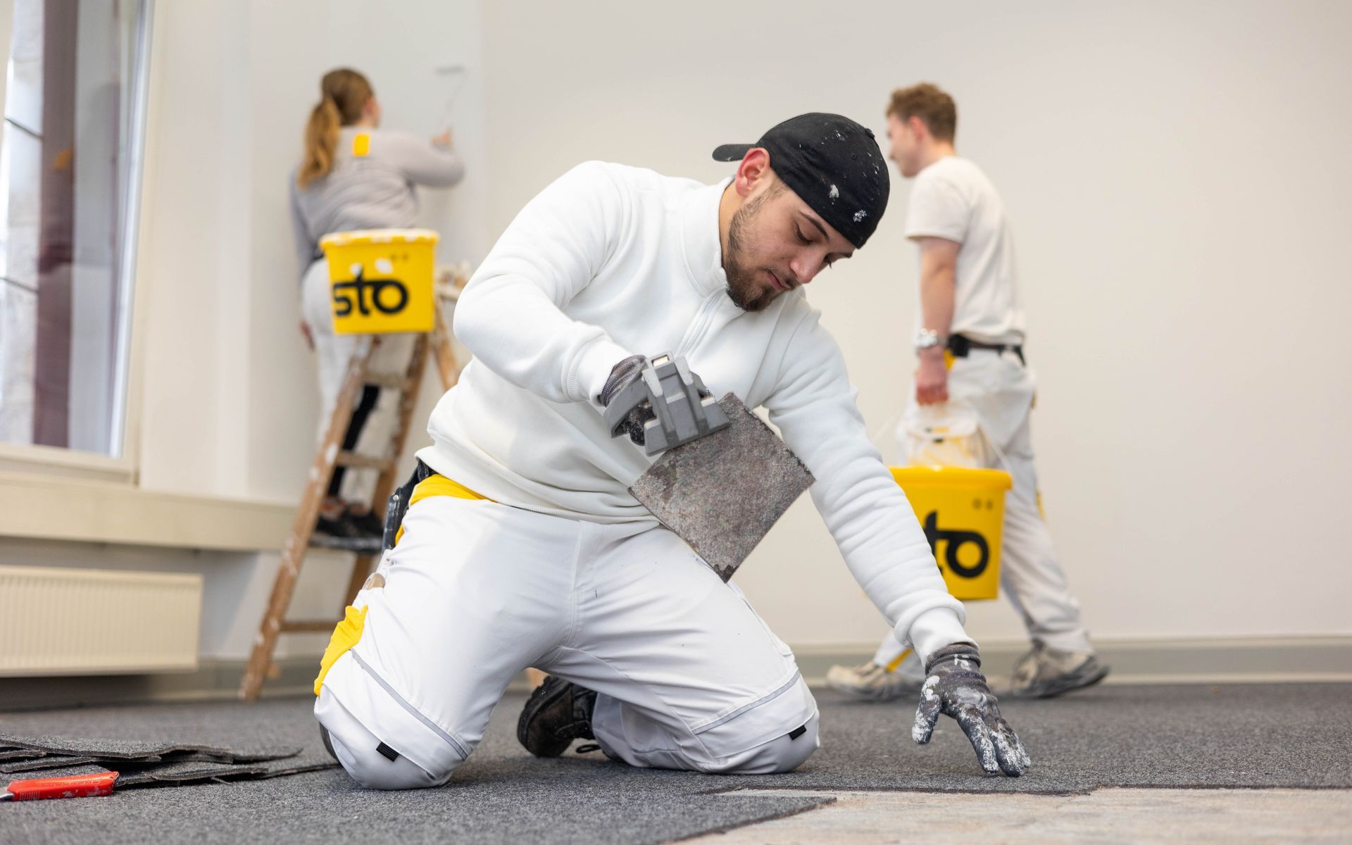 Ein Mann kniet auf dem Boden, während im Hintergrund eine Frau auf einer Leiter steht.