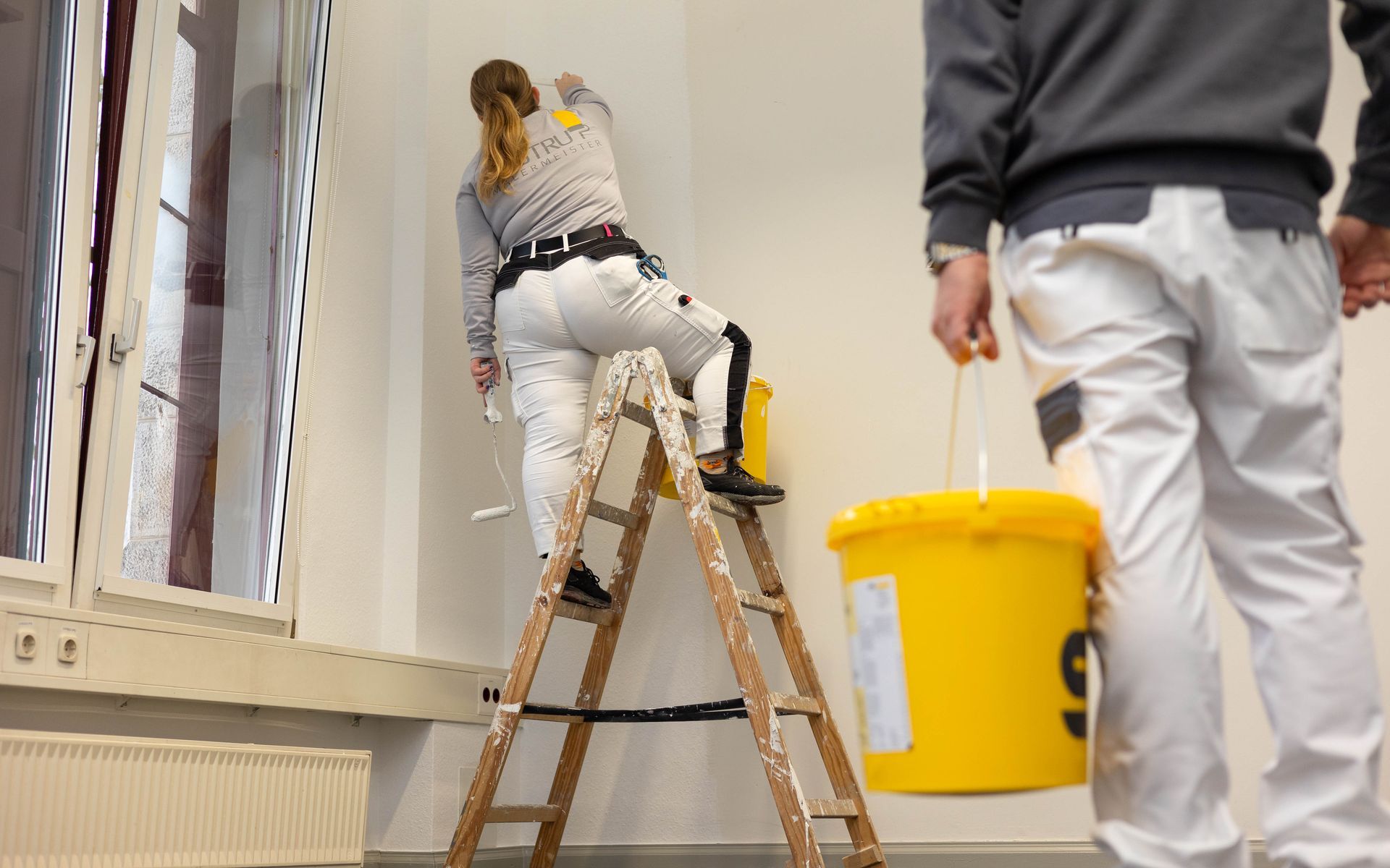 Eine Frau streicht auf einer Leiter eine Wand, während ein Mann einen gelben Eimer hält.