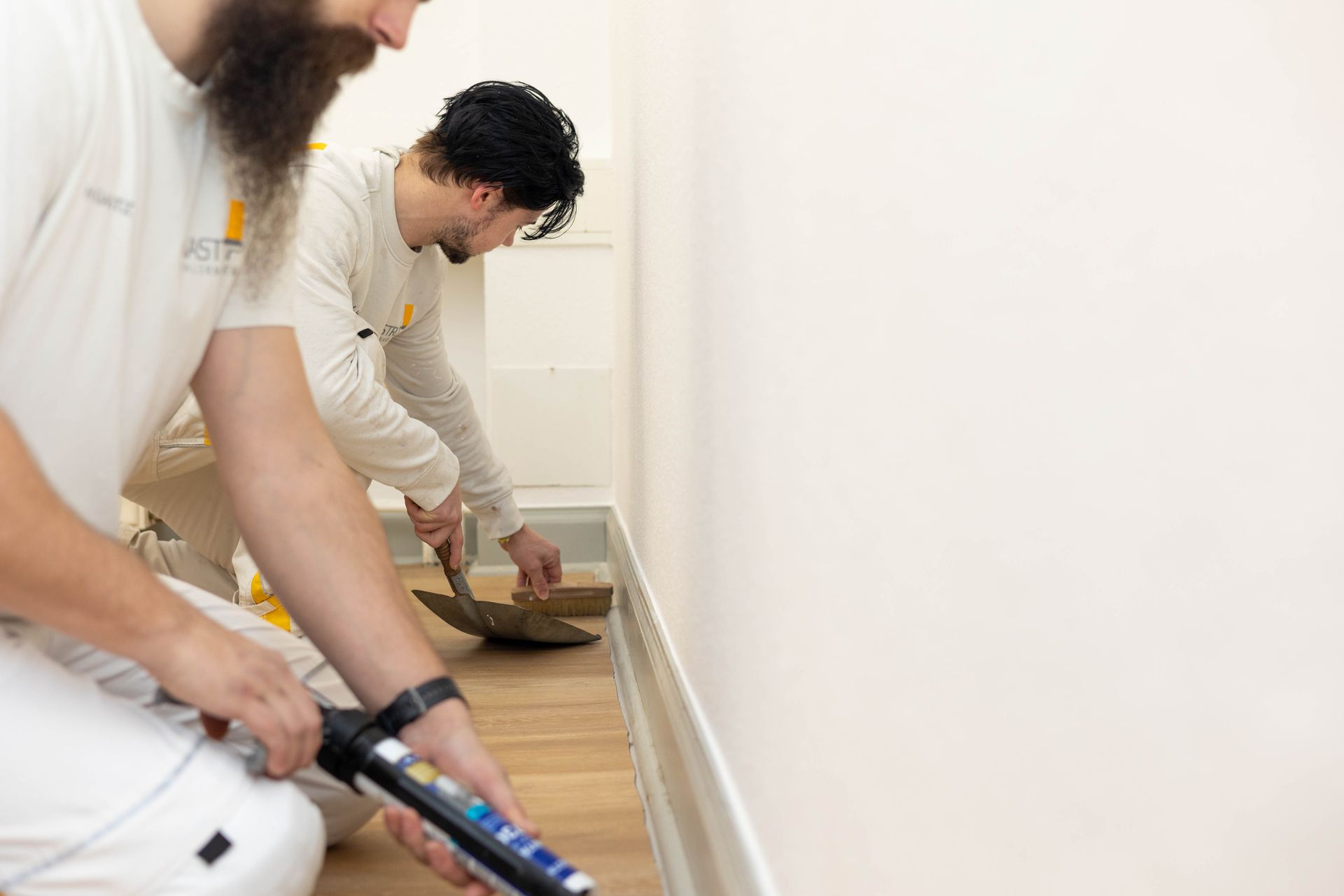 Zwei Männer knien in einem Flur und einer von ihnen hält eine Flasche Klebstoff in der Hand.
