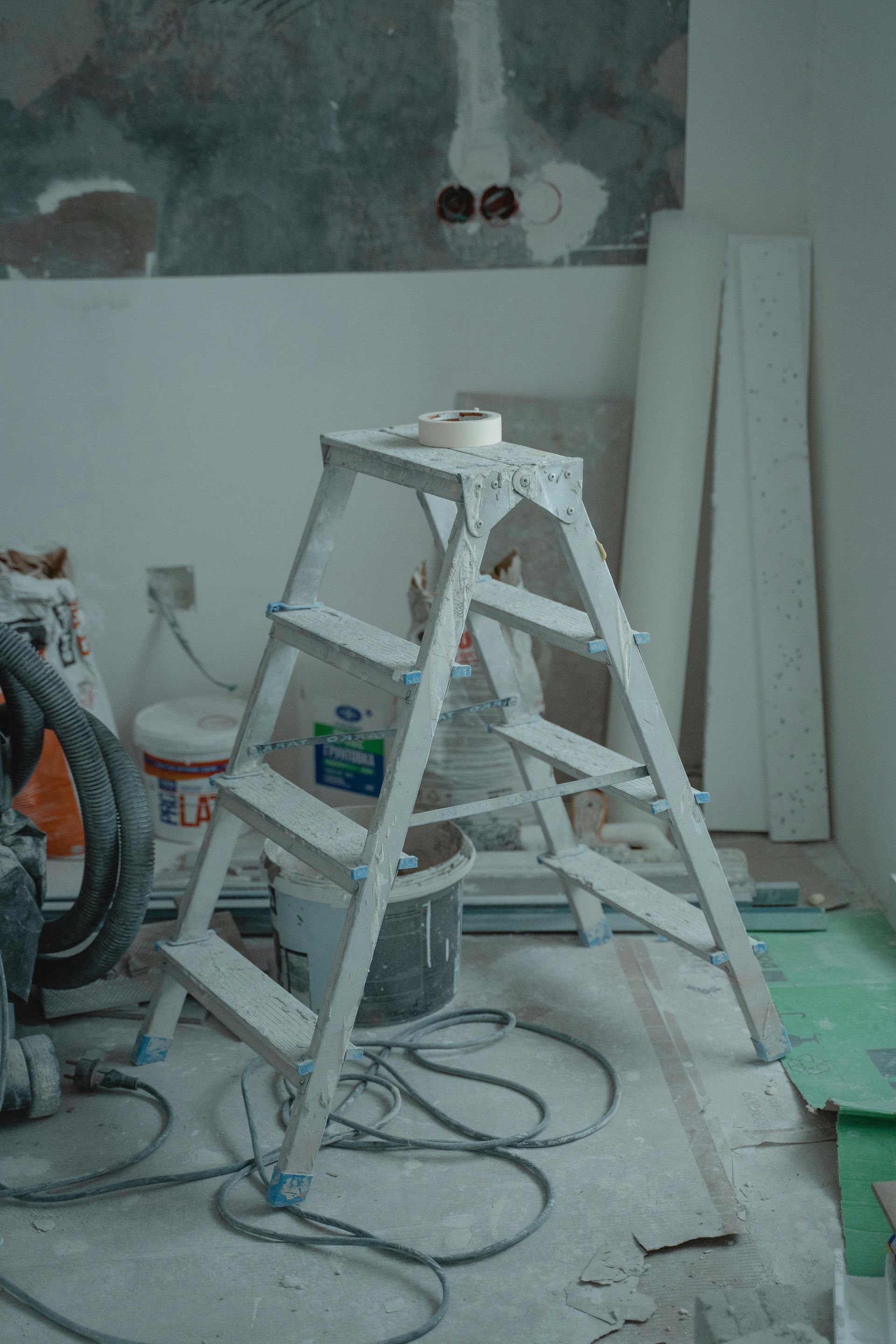 Échelle à tréteaux peinte dans une pièce de rénovation poussiéreuse, avec des outils, des câbles et des planches appuyés contre le mur.
