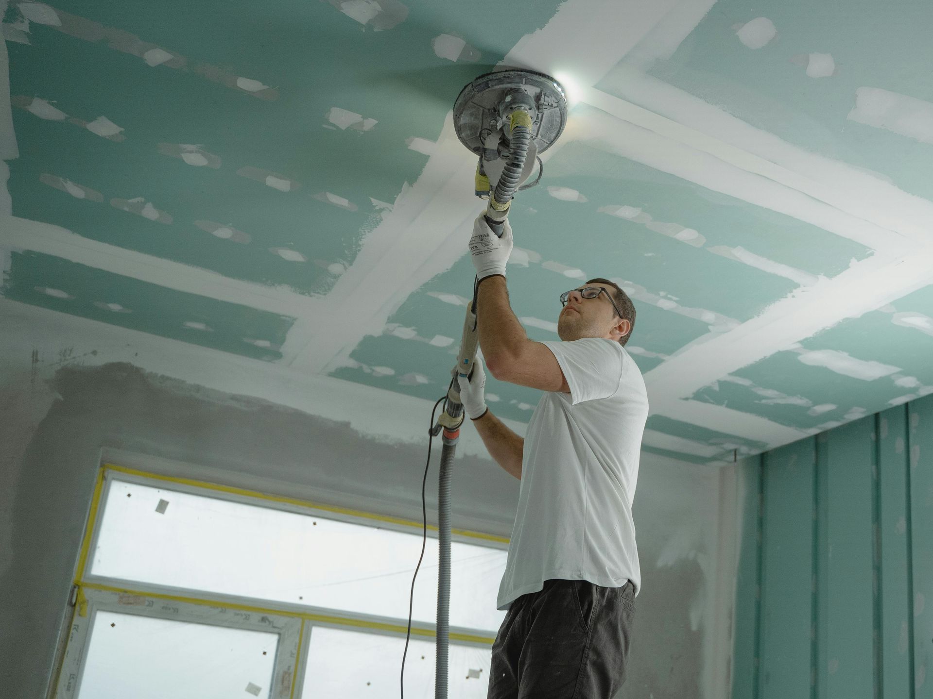 Un ouvrier ponce un plafond avec une ponceuse électrique dans une pièce en rénovation