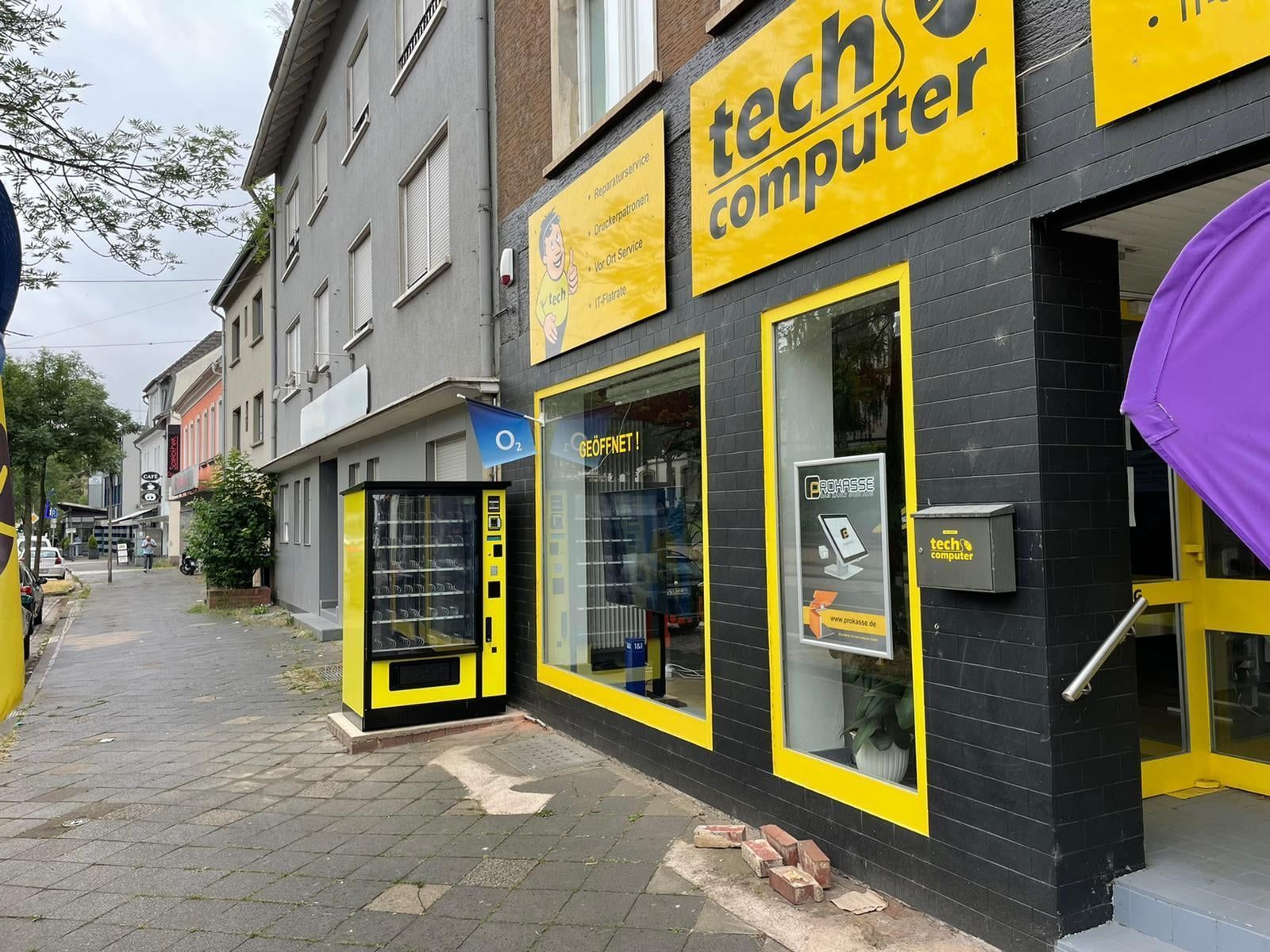 Gelb-schwarzes Computergeschäft mit Schaufenster und Hinweisschild auf dem Bürgersteig in einer schmalen Kopfsteinpflasterstraße.