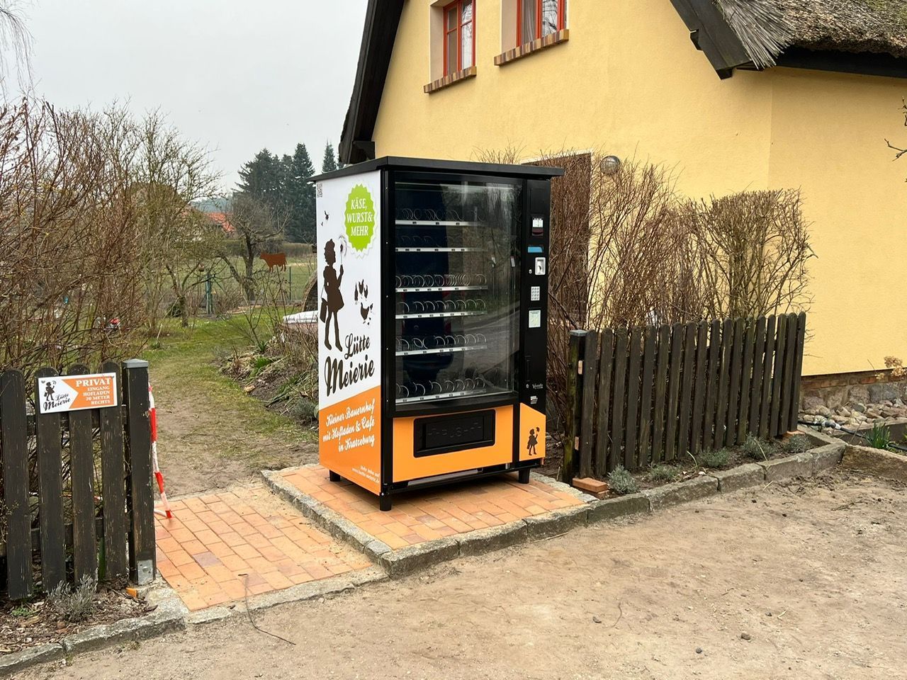 Ein Verkaufsautomat im Freien steht neben einem gelben Haus und einem Holzzaun, dahinter befinden sich ein gepflasterter Weg und ein Garten.