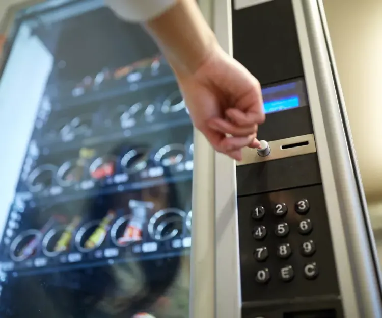 Eine Hand gibt einen Code auf dem Tastenfeld eines Verkaufsautomaten neben einem Getränkedisplay ein.