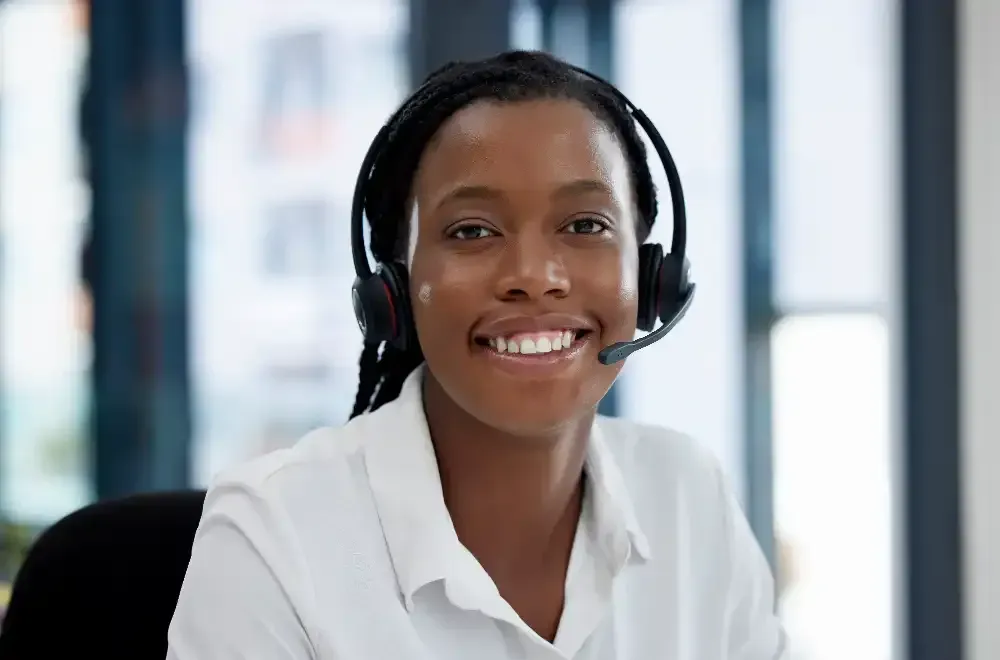 A woman is wearing a headset and smiling while sitting at a desk.