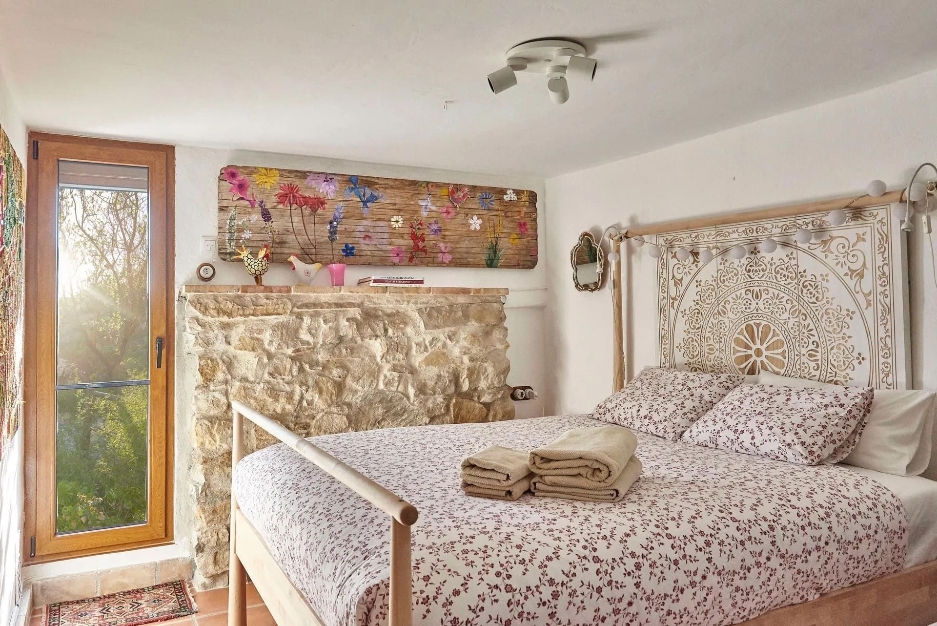 Dormitorio con cama, pared de piedra, obras de arte coloridas y una ventana con luz solar.