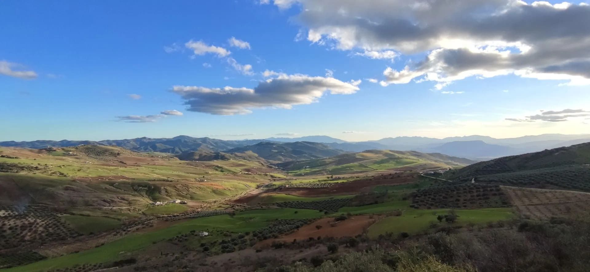 Colinas verdes onduladas bajo un cielo azul con nubes blancas; el sol brilla en el paisaje.