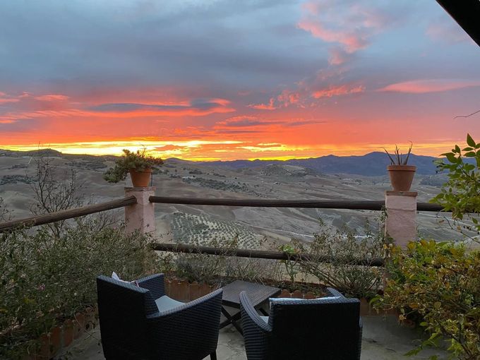 Atardecer sobre un vasto paisaje visto desde un patio con asientos y barandilla. Cielo naranja y rojo brillante.