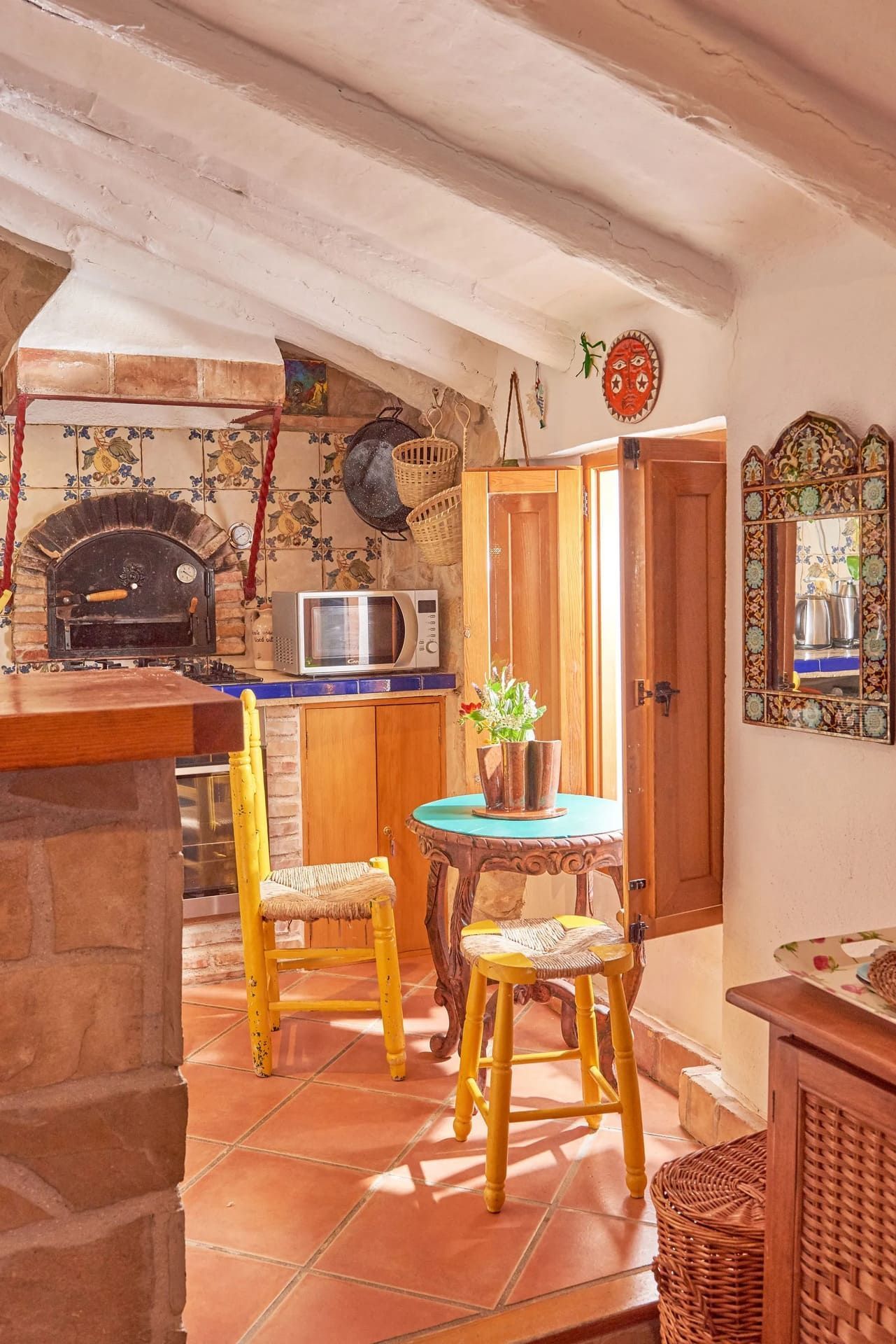 Cocina rústica con horno de ladrillo, sillas amarillas y pequeña mesa redonda.