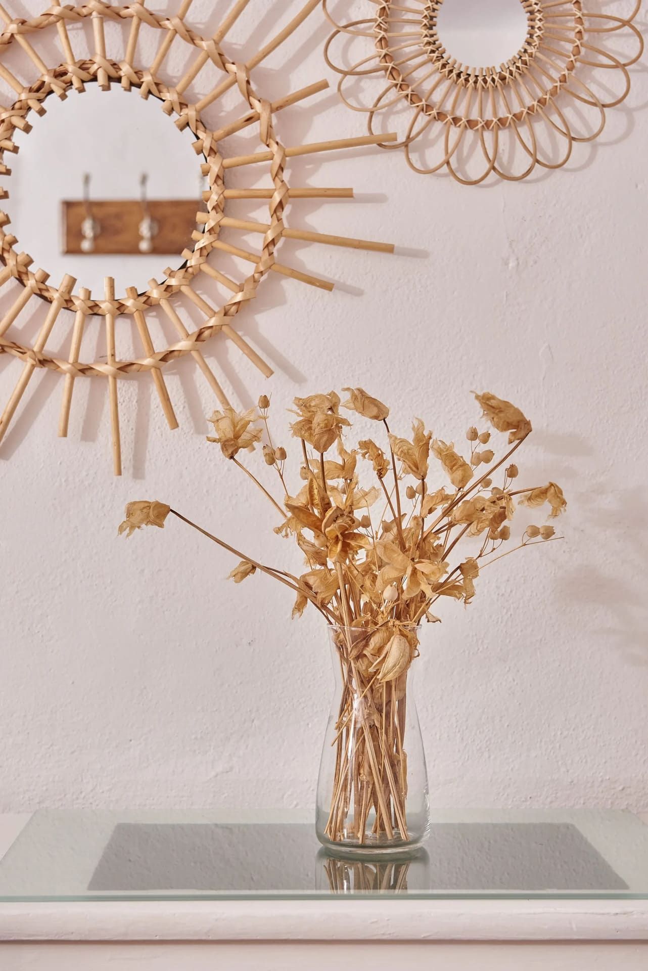 Flores secas en un jarrón de cristal sobre un estante, con espejos de ratán con forma de sol y un perchero de madera.