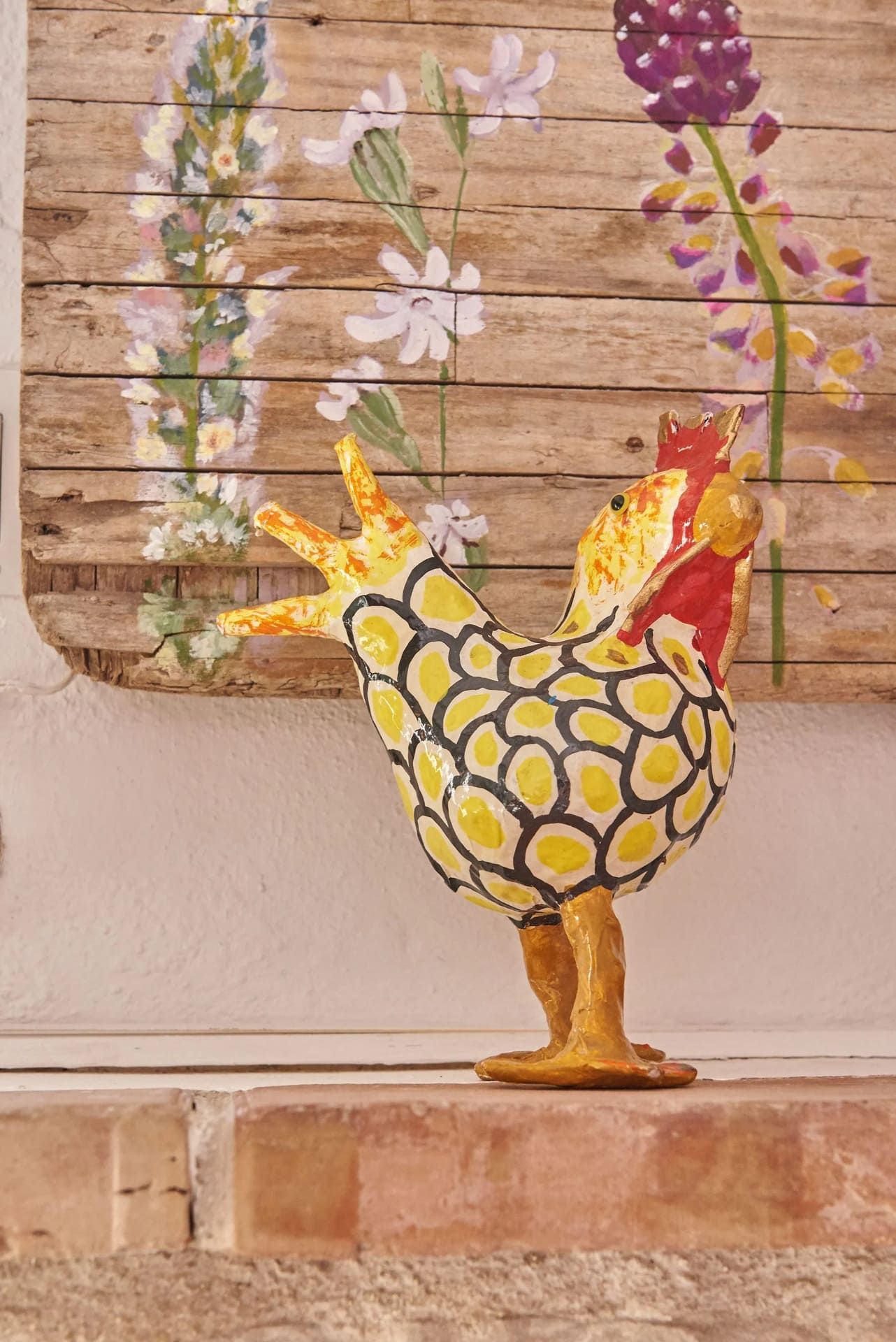 Escultura de gallo con manchas amarillas y negras sobre una repisa de ladrillo, con pintura floral de fondo.
