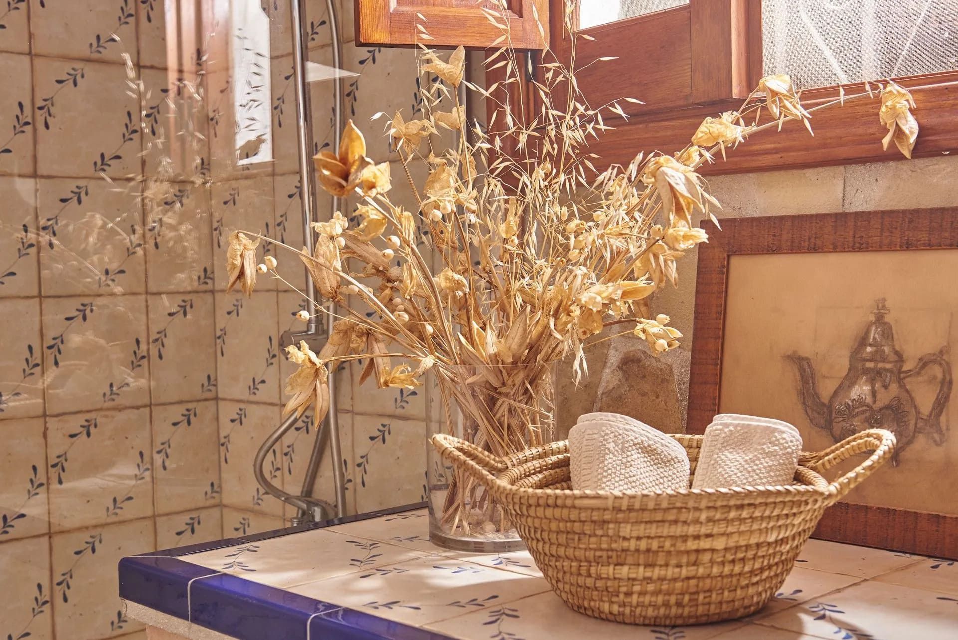 Cesta de toallas y flores secas sobre un mostrador, junto a una ventana en un baño.
