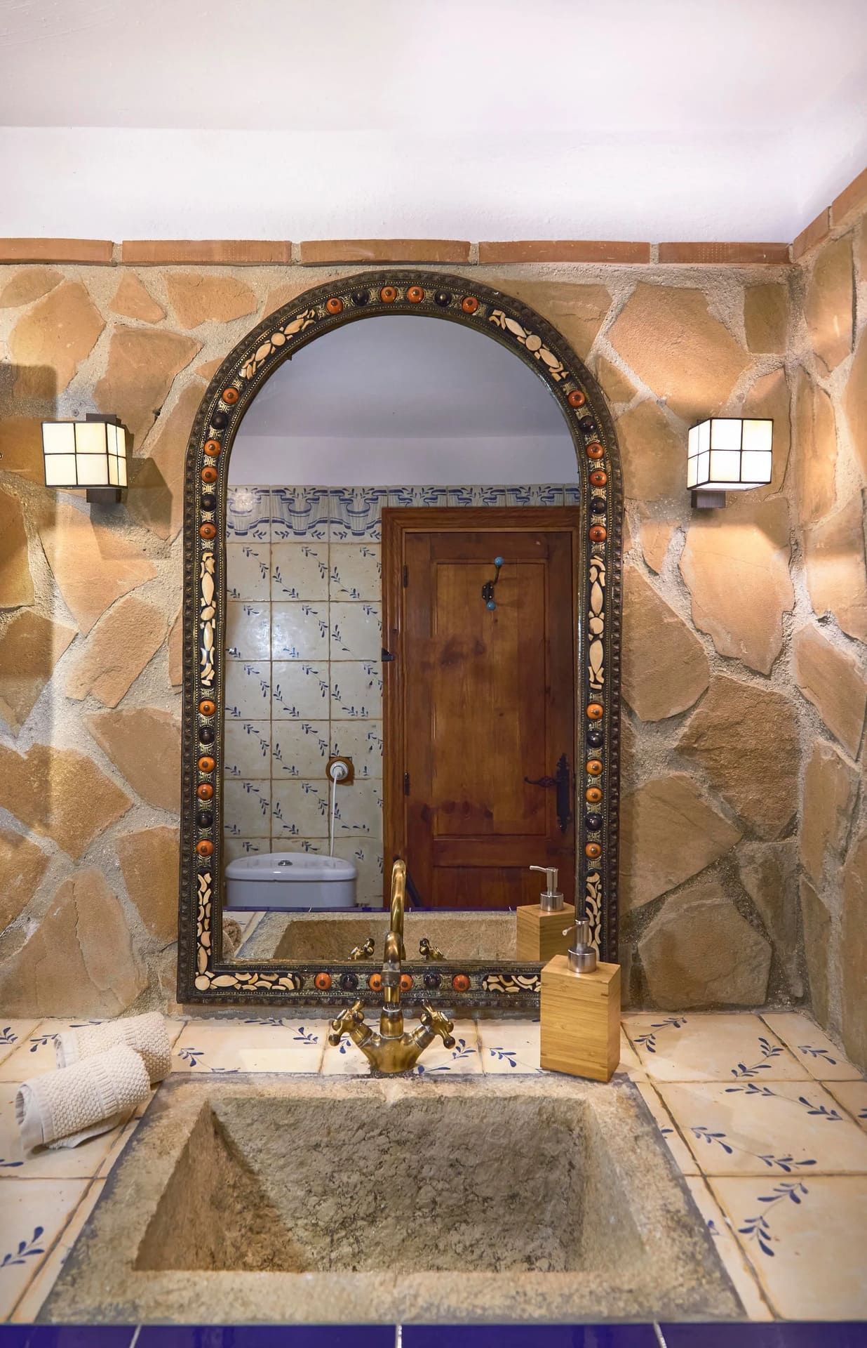 Baño con paredes de piedra, gran espejo arqueado, lavabo de piedra y encimera de azulejos azules.