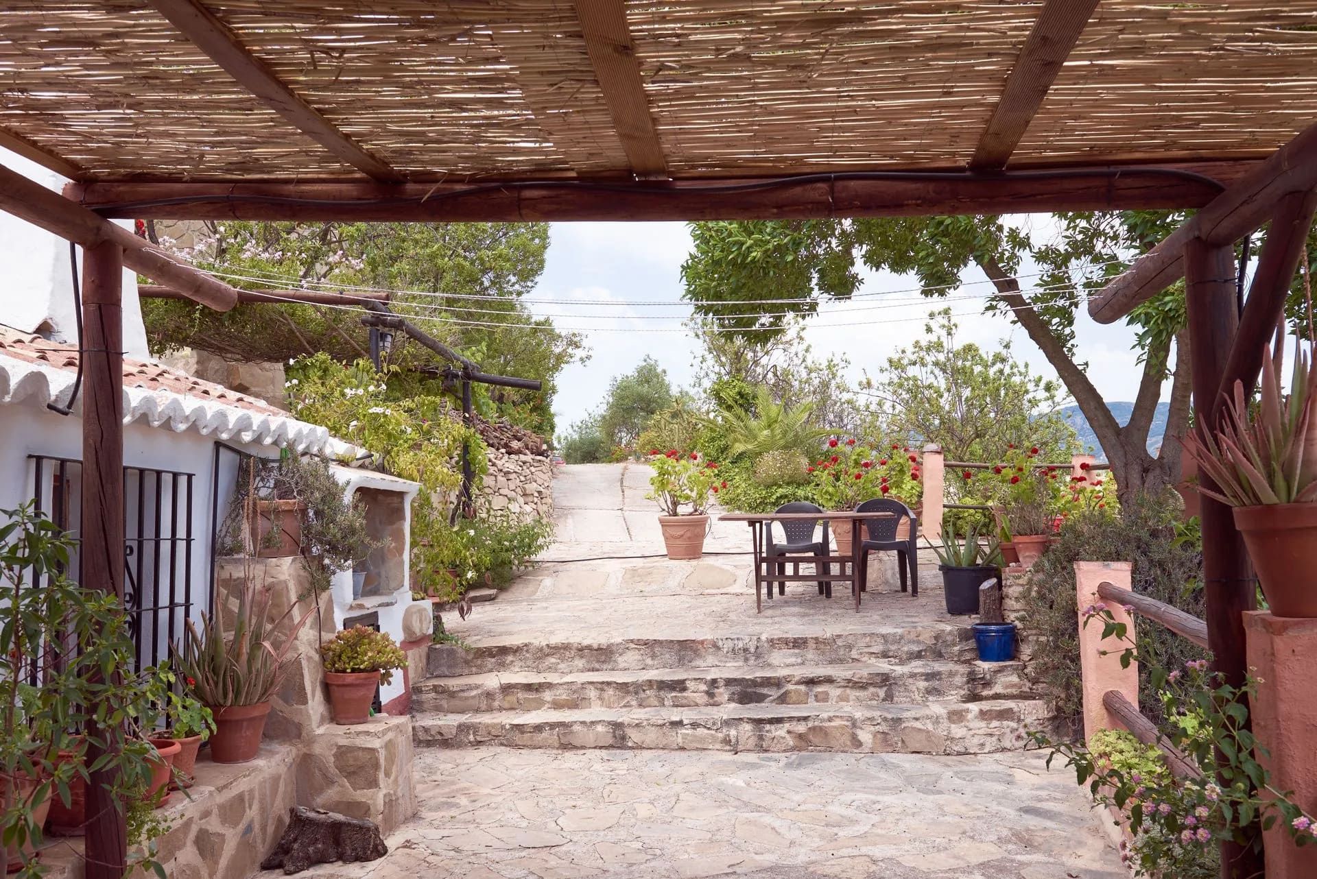 Unos escalones de piedra conducen a una mesa y sillas, a la sombra de una pérgola. Plantas y una pequeña casa bordean el camino.