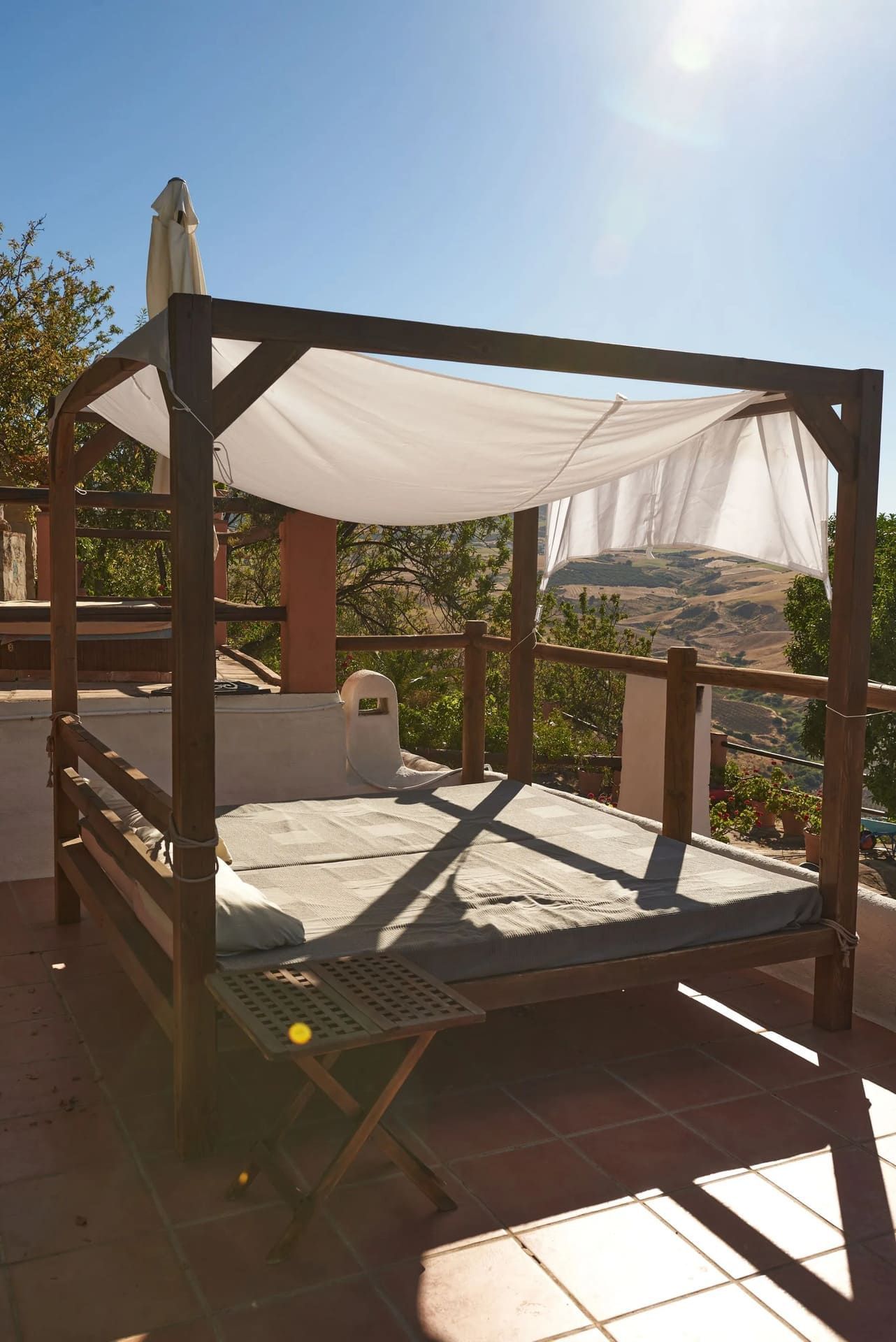Cama con dosel de madera y tela blanca sobre un patio de baldosas, con vistas a un paisaje soleado.