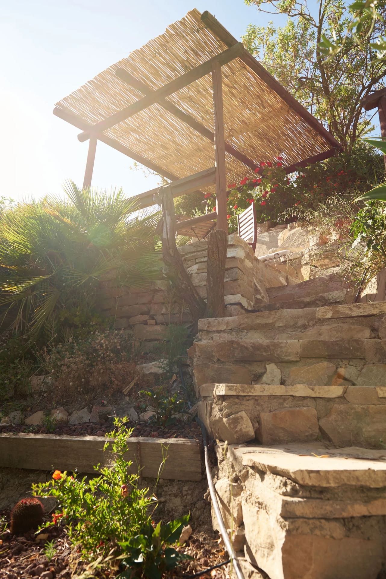 Unos escalones de piedra conducen a una estructura con techo de paja, ubicada en un jardín soleado.