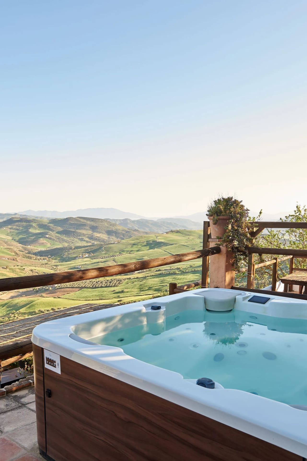Puesta de sol sobre colinas verdes con nubes rosas y naranjas, vista desde detrás de una barandilla con flores.