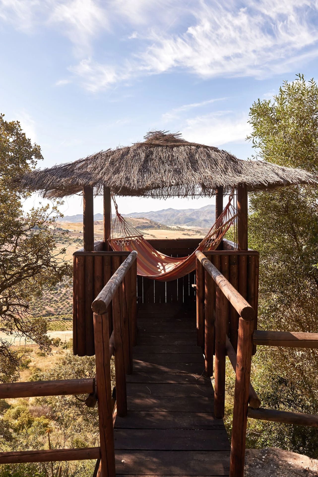 Cenador de madera con techo de paja y hamaca, puente que conduce al mismo, ubicado en un paisaje natural y soleado.