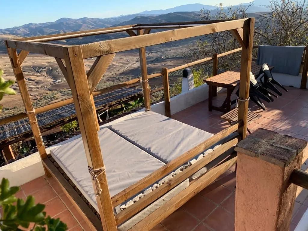 Tumbona de madera para exterior con cojines en terraza con vistas a un valle, día soleado.