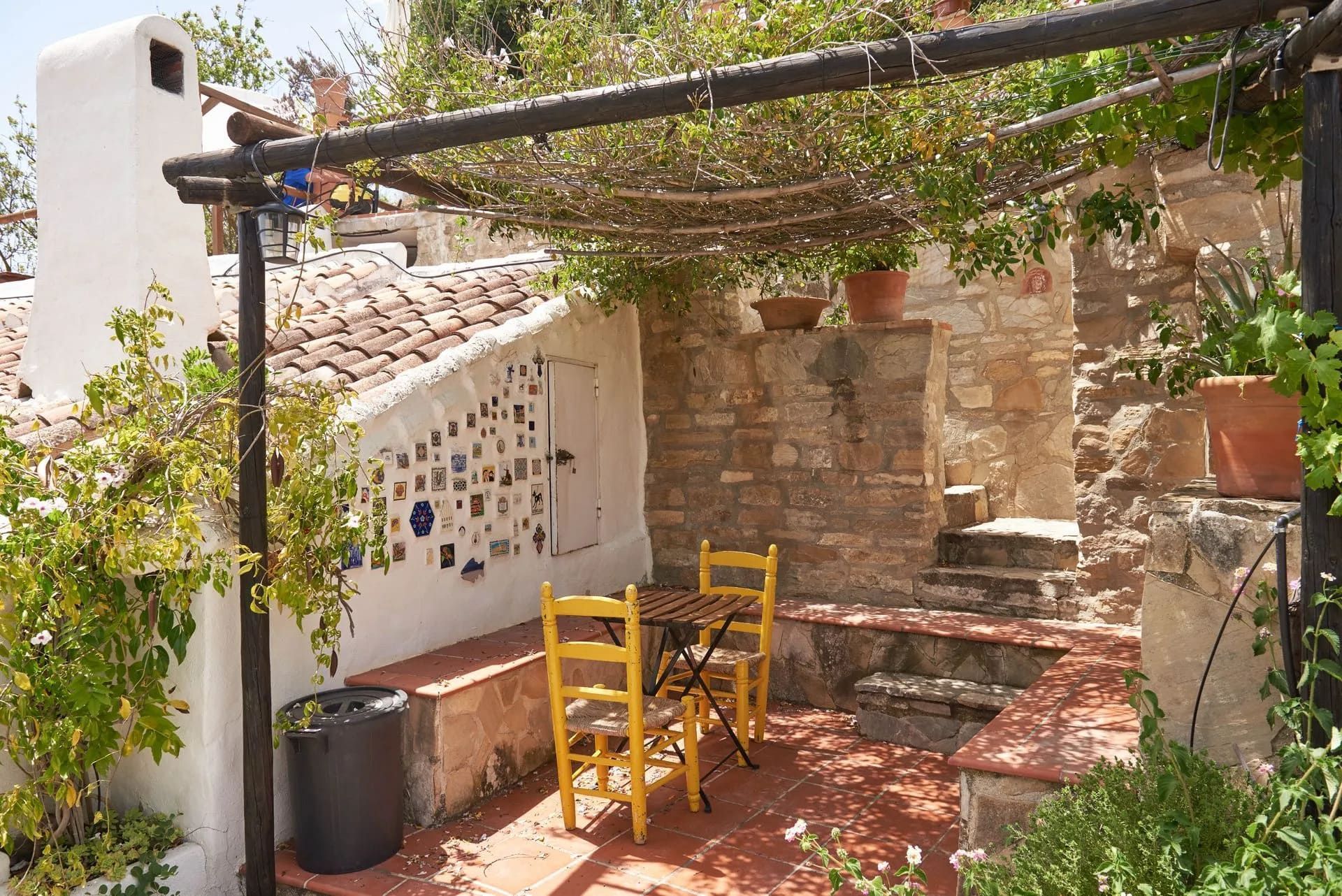 Patio al aire libre con sillas amarillas, sombreado por enredaderas, paredes de ladrillo y escaleras.