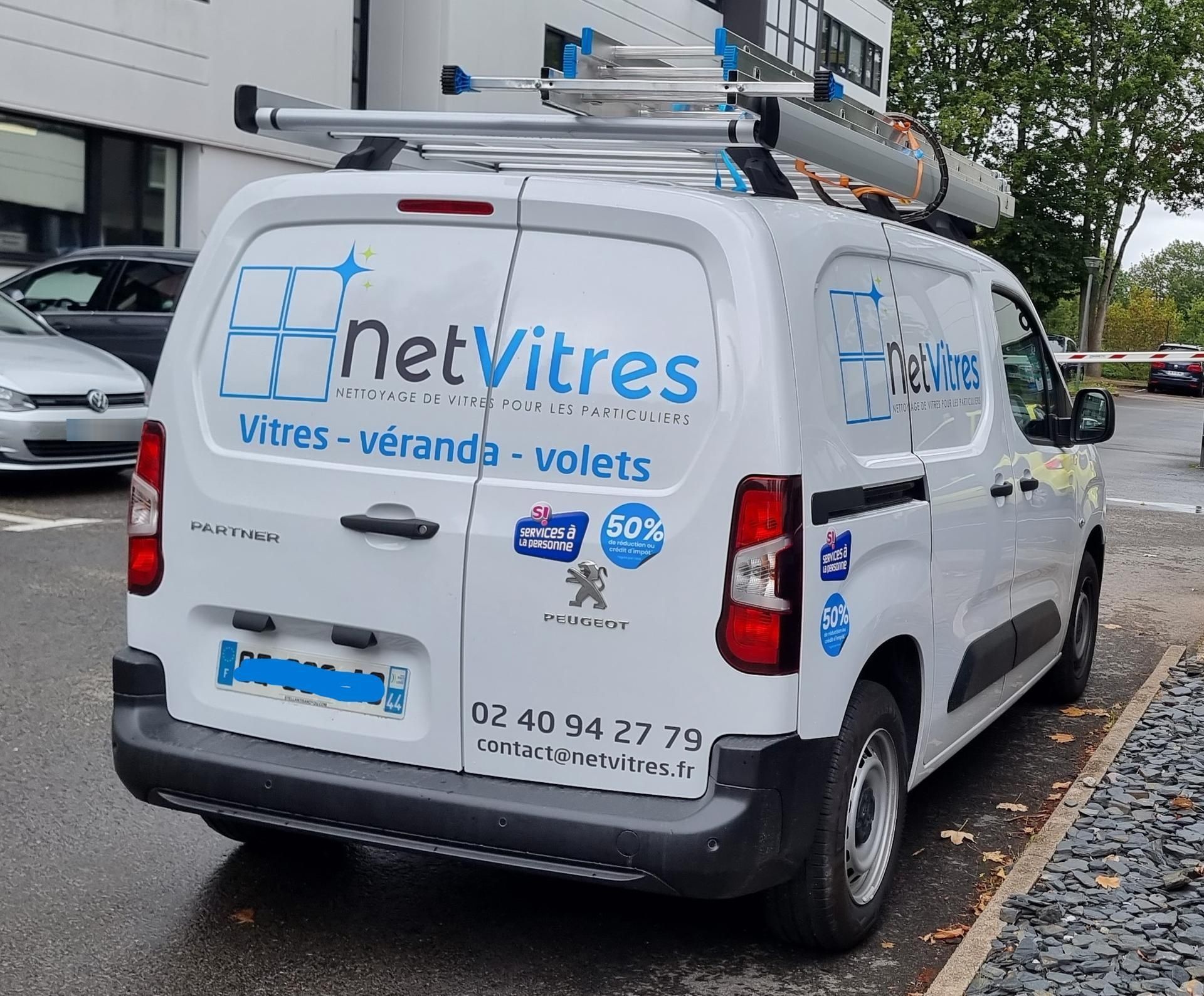 Fourgonnette de service blanche NetVitres stationnée dans la rue, avec logo bleu de l'entreprise et porte-échelle de toit