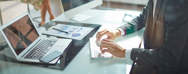 Una persona sentada en su escritorio con una computadora portátil y una taza de café, revisando documentos en una oficina luminosa.