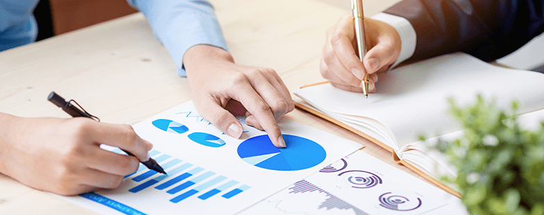 Manos revisando gráficos y tablas azules sobre un escritorio con bolígrafos y papeles.