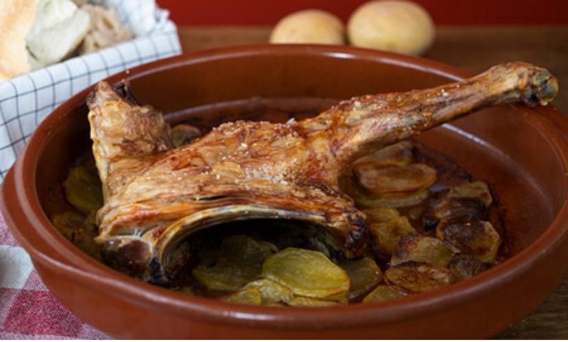 Cordero asado con patatas en rodajas en una fuente de barro marrón, con una cesta de pan forrada con un paño al fondo.
