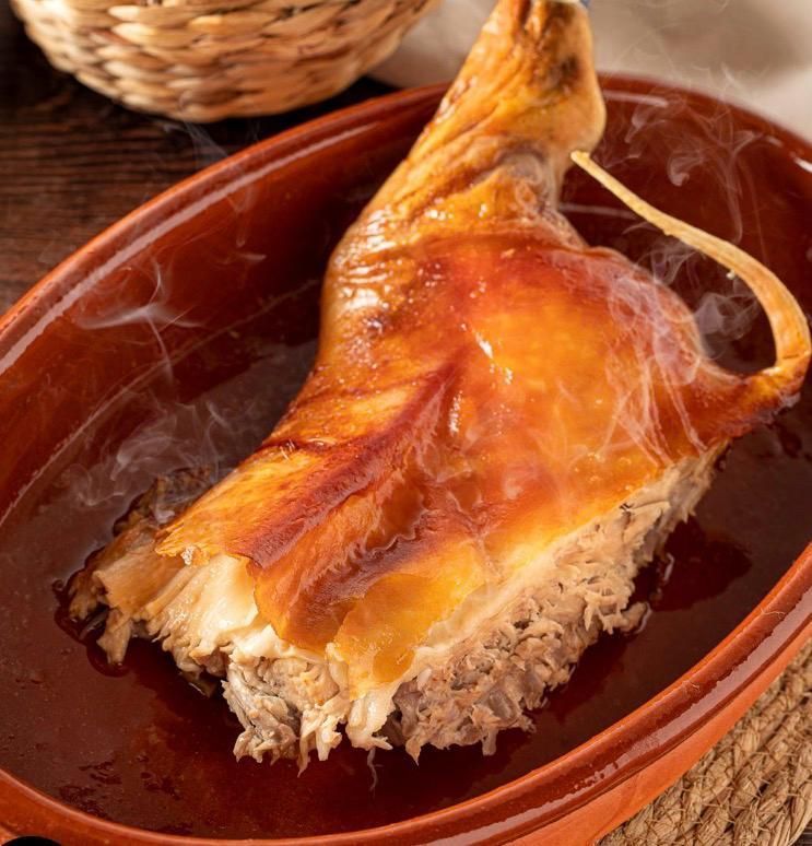 Una pierna de cochinillo asada, dorada y crujiente, servida en una fuente de barro humeante.