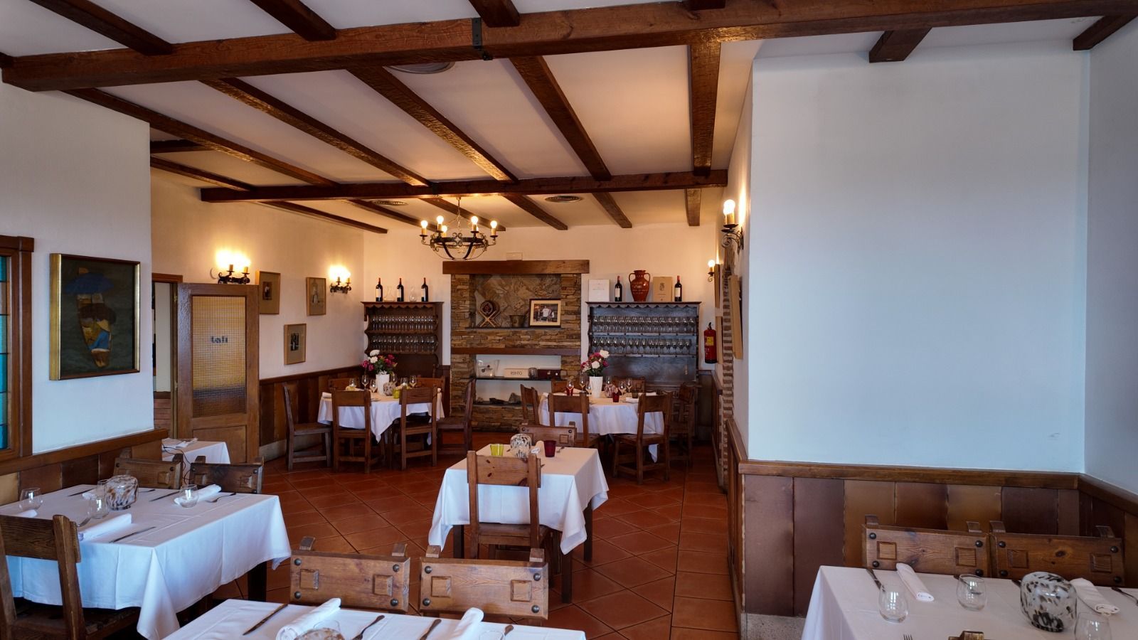 Comedor rústico con techos con vigas de madera, mesas con manteles blancos y una chimenea de piedra al fondo.