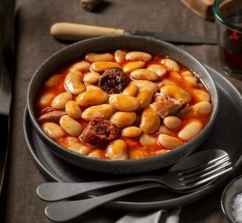 Un plato de guiso de alubias con trozos de salchicha oscura, servido en un plato hondo sobre una mesa 