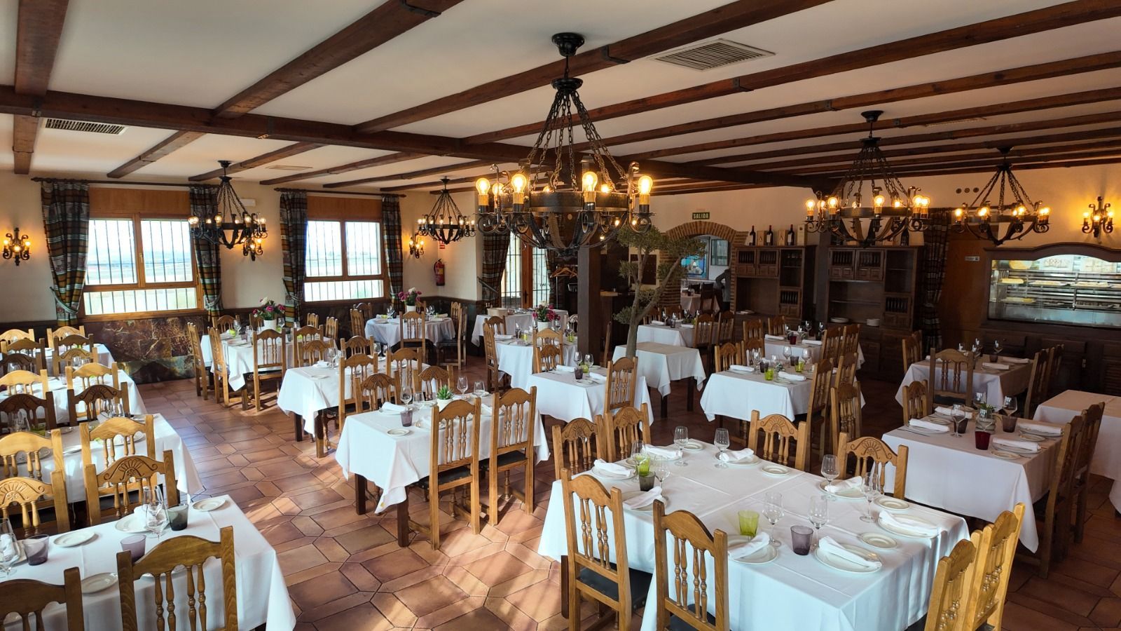 Un restaurante rústico y espacioso con vigas de madera, lámparas de araña y varias mesas vestidas con manteles blancos.