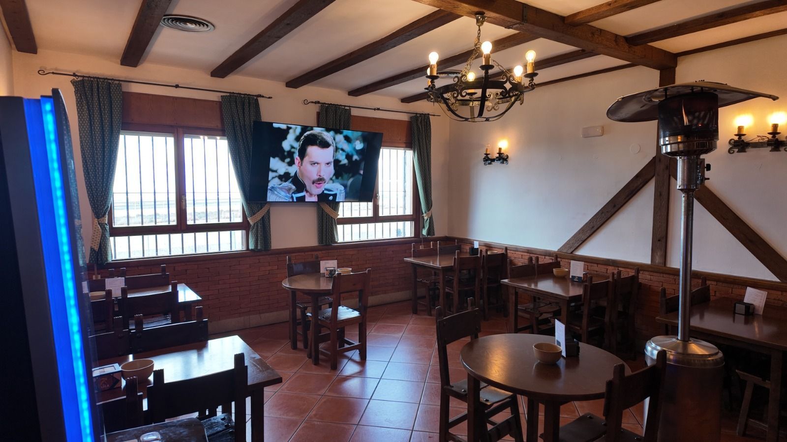 Interior de un pub con mesas y sillas de madera, un televisor que muestra a un músico, una lámpara de araña 