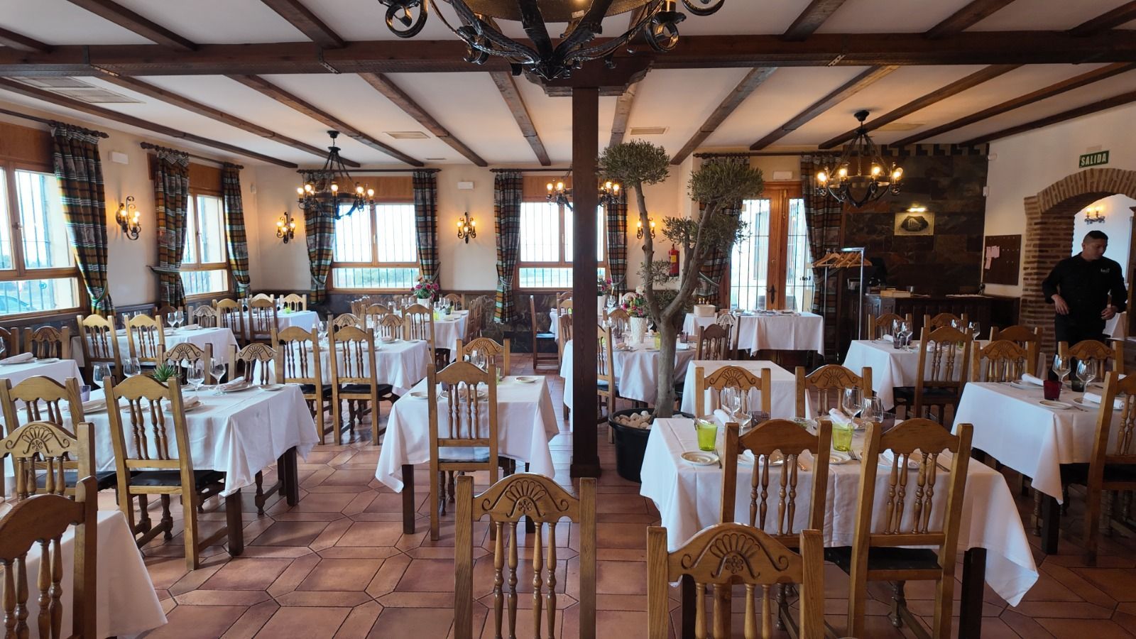 Interior rústico del restaurante con vigas de madera, suelos de baldosas, varias mesas con manteles blancos 