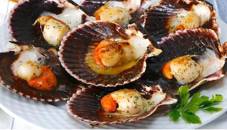 Vieiras cocidas en sus conchas oscuras y estriadas, sazonadas con hierbas y servidas en un plato 