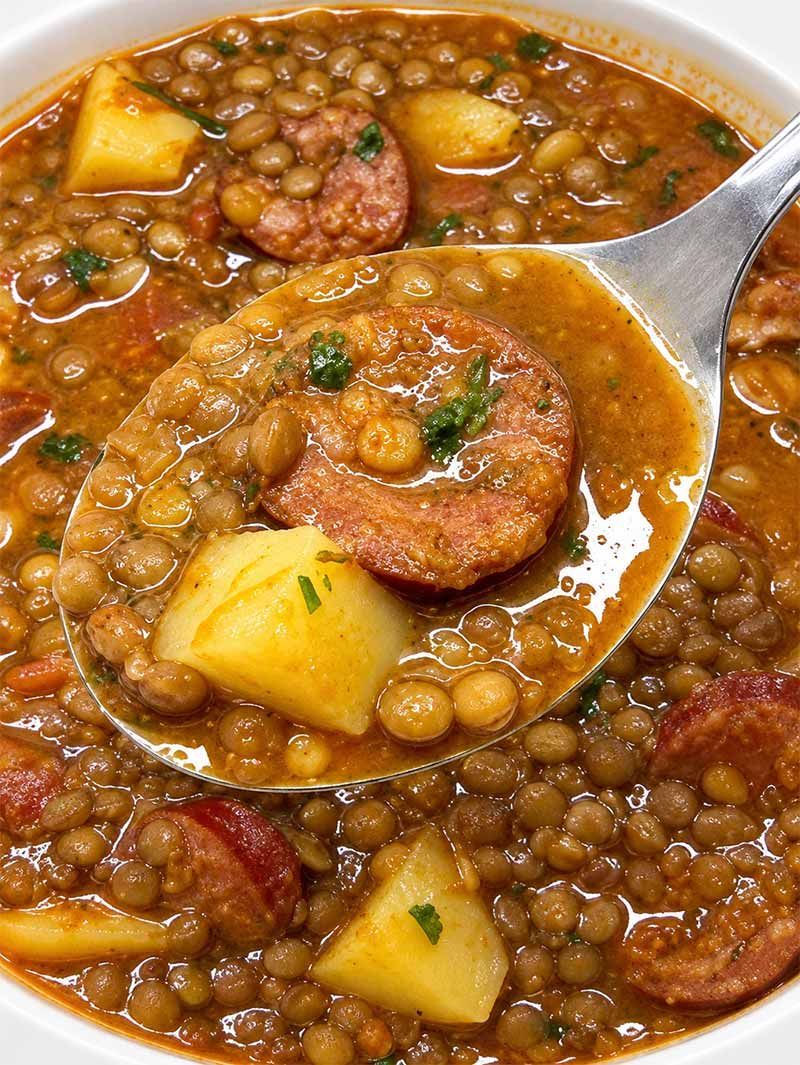 Cucharón sirviendo un guiso de lentejas con salchicha y trozos de patata en un cuenco