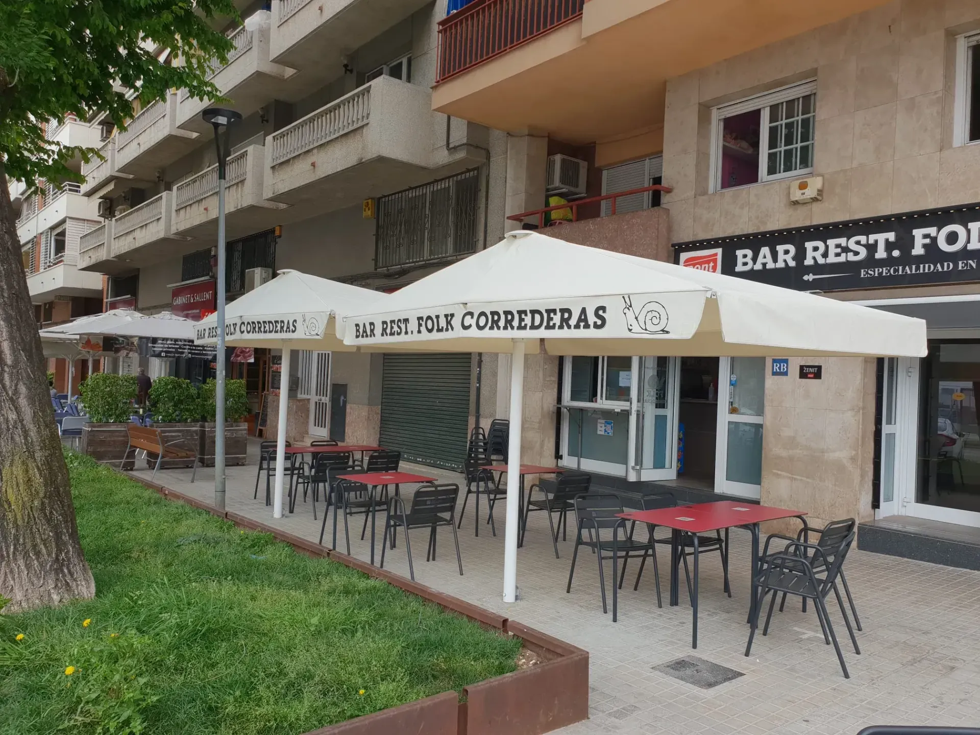 Mesas y sillas bajo sombrillas frente a un edificio que dice bar rest