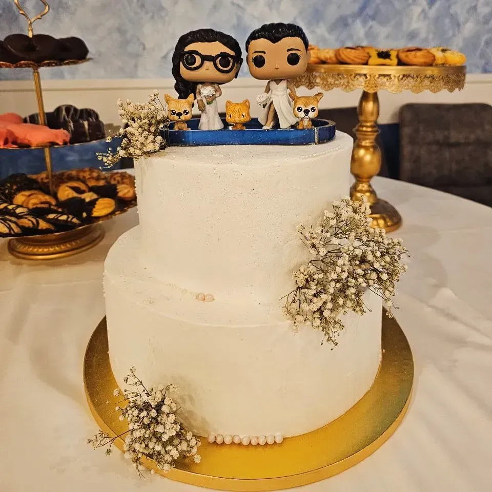 Tarta nupcial blanca de dos pisos con figuras de la pareja y flores. Soportes dorados para tartas sostienen los postres en el lugar de la celebración.