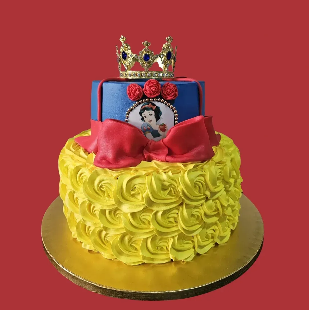 Tarta de dos pisos con glaseado amarillo, un lazo rojo y la imagen de Blancanieves. Coronada con una capa azul y una corona dorada.