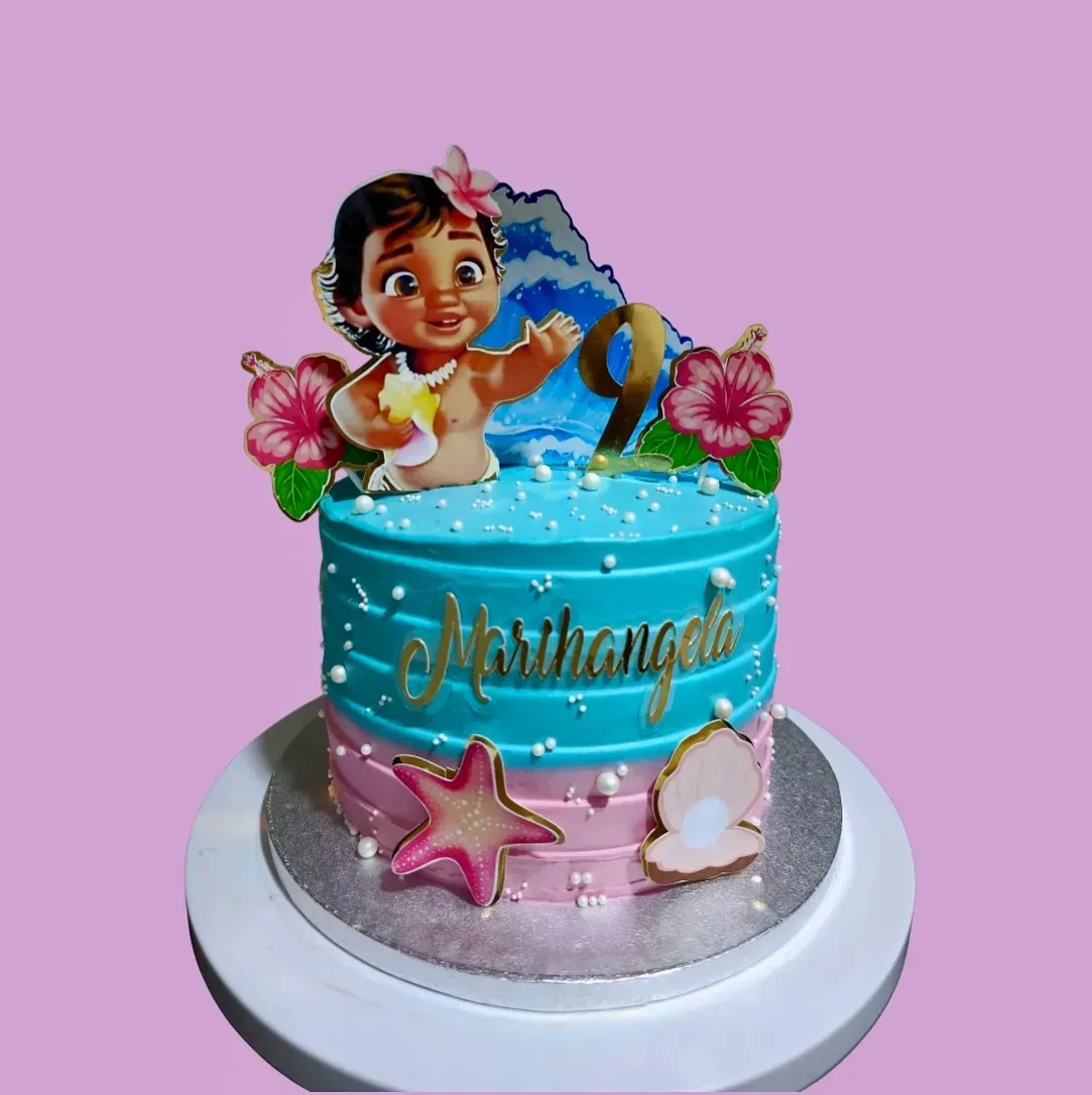 Tarta de cumpleaños inspirada en la Bebé Moana, con glaseado azul océano y rosa, decorada con elementos comestibles y un adorno.