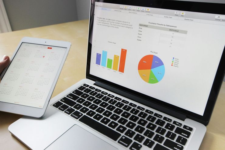 ordenador abierto con gráficos en el y una persona sujetando al lado una tablet de color blanco con más información 