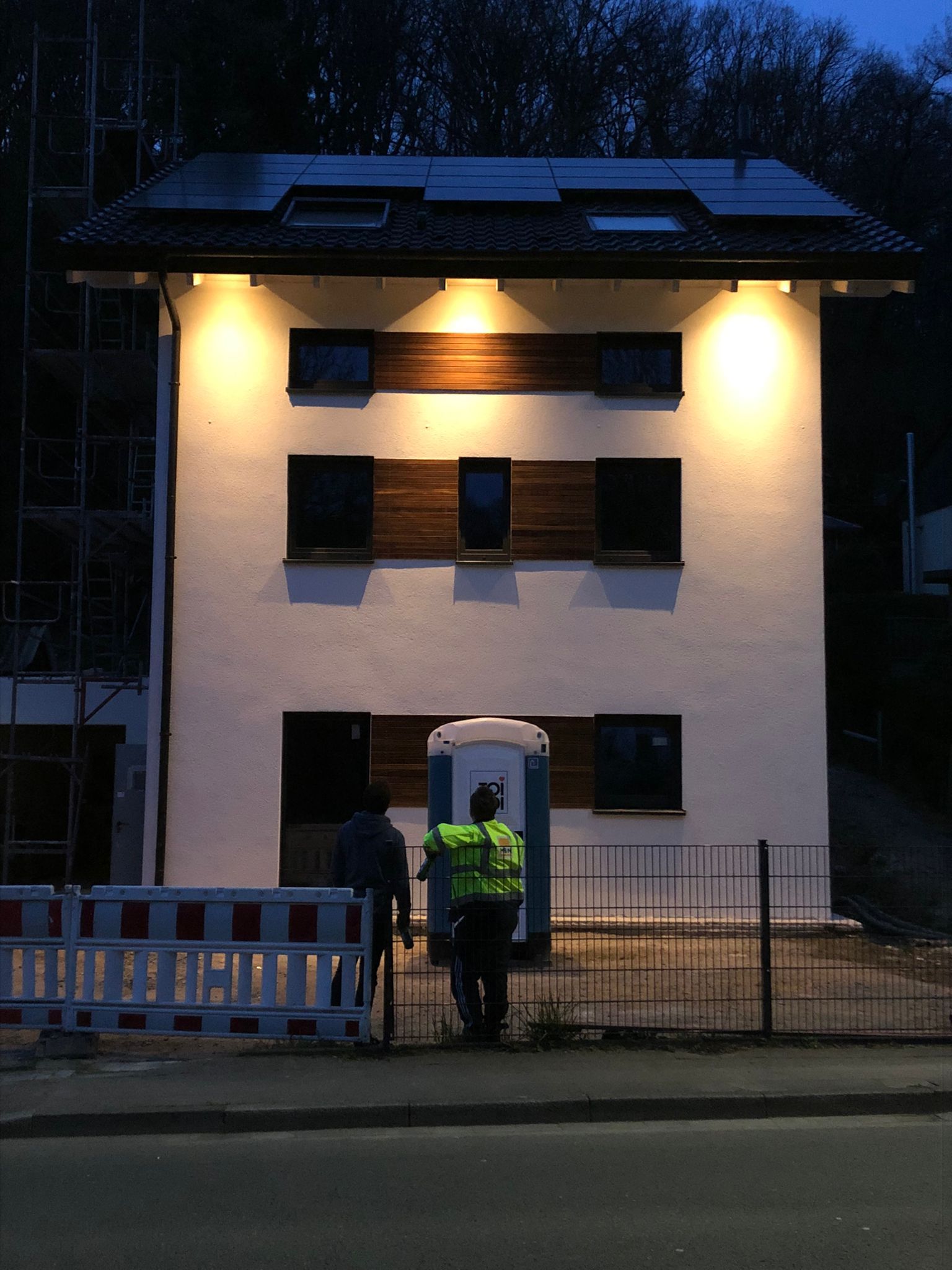 Zweistöckiges weißes Gebäude, nachts beleuchtet, mit Solarzellen auf dem Dach. Bauarbeiter vor dem Zaun.