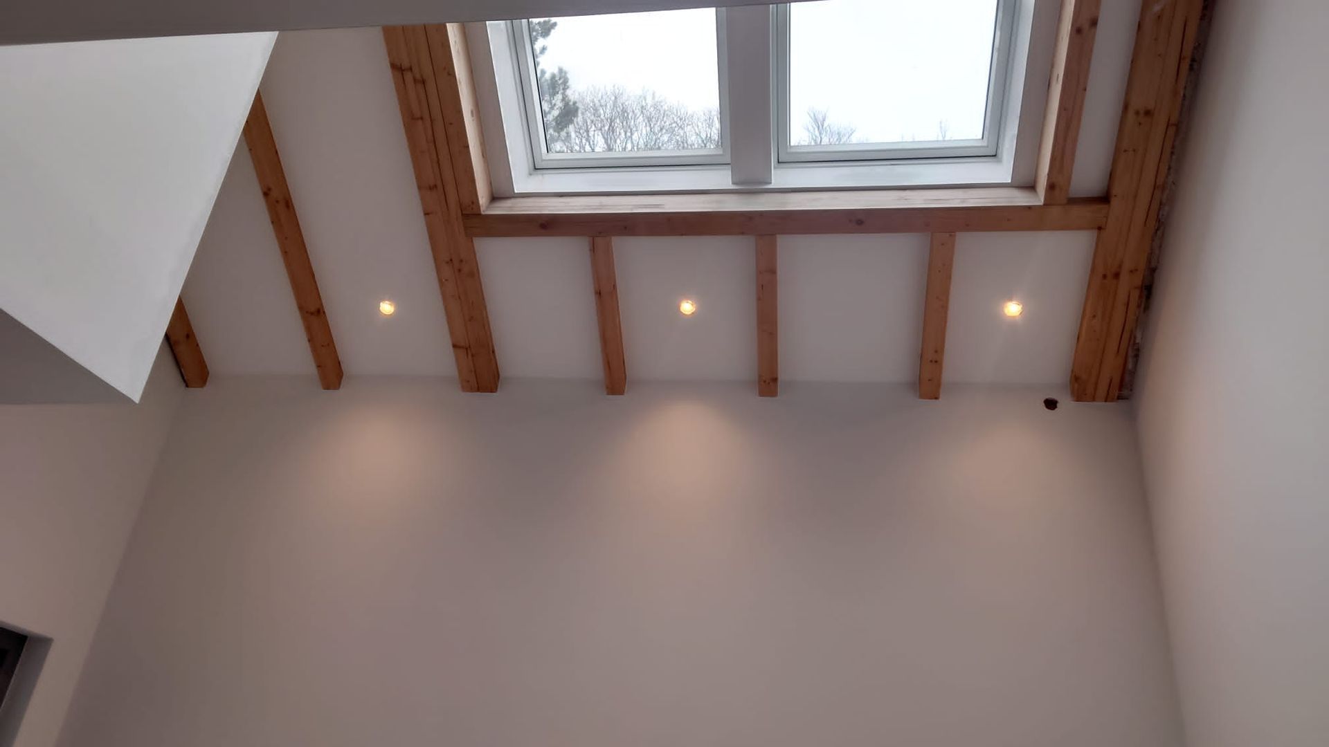 Raum mit hoher Decke, freiliegenden Holzbalken, Oberlicht und Einbauleuchten, die die weißen Wände erhellen.