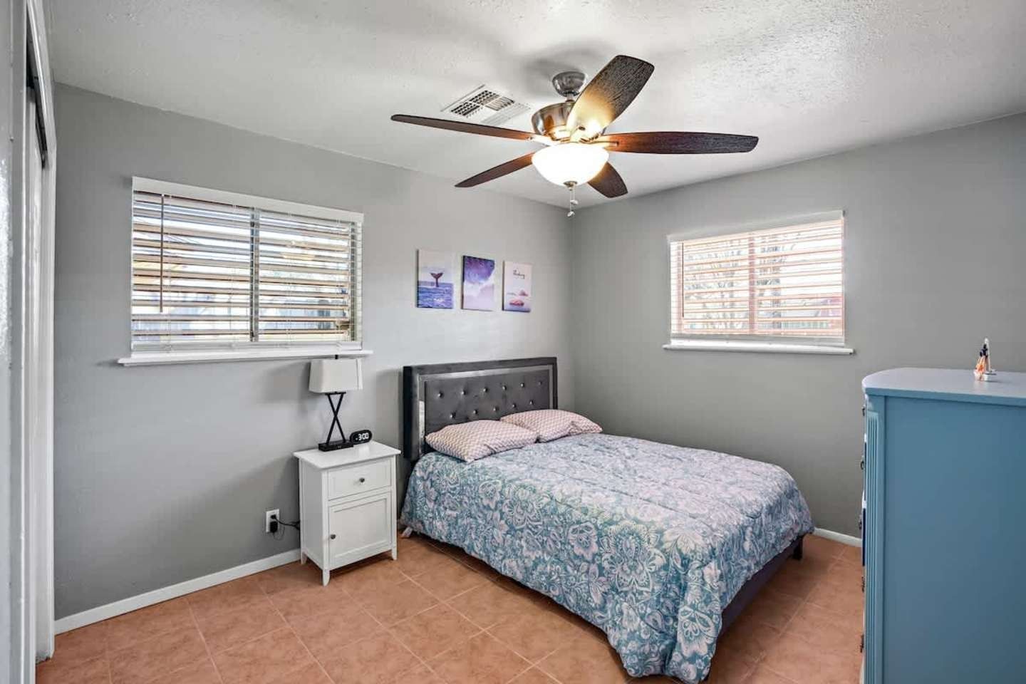 Small bedroom with gray walls, a bed, nightstand, ceiling fan, and two windows with blinds.