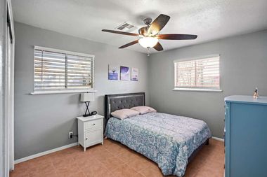 Small bedroom with gray walls, a bed, nightstand, ceiling fan, and two windows with blinds.