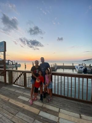Family of four posing on a wooden boardwalk at sunset by the water.