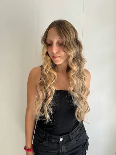 Mujer con cabello rubio largo y ondulado, vestida con una camiseta sin mangas negra y jeans, mirando hacia abajo.