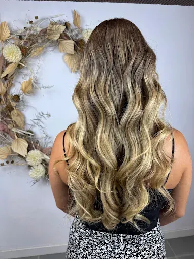 Mujer con cabello largo, ondulado y estilo ombré, de pie frente a una pared blanca con una corona de flores.
