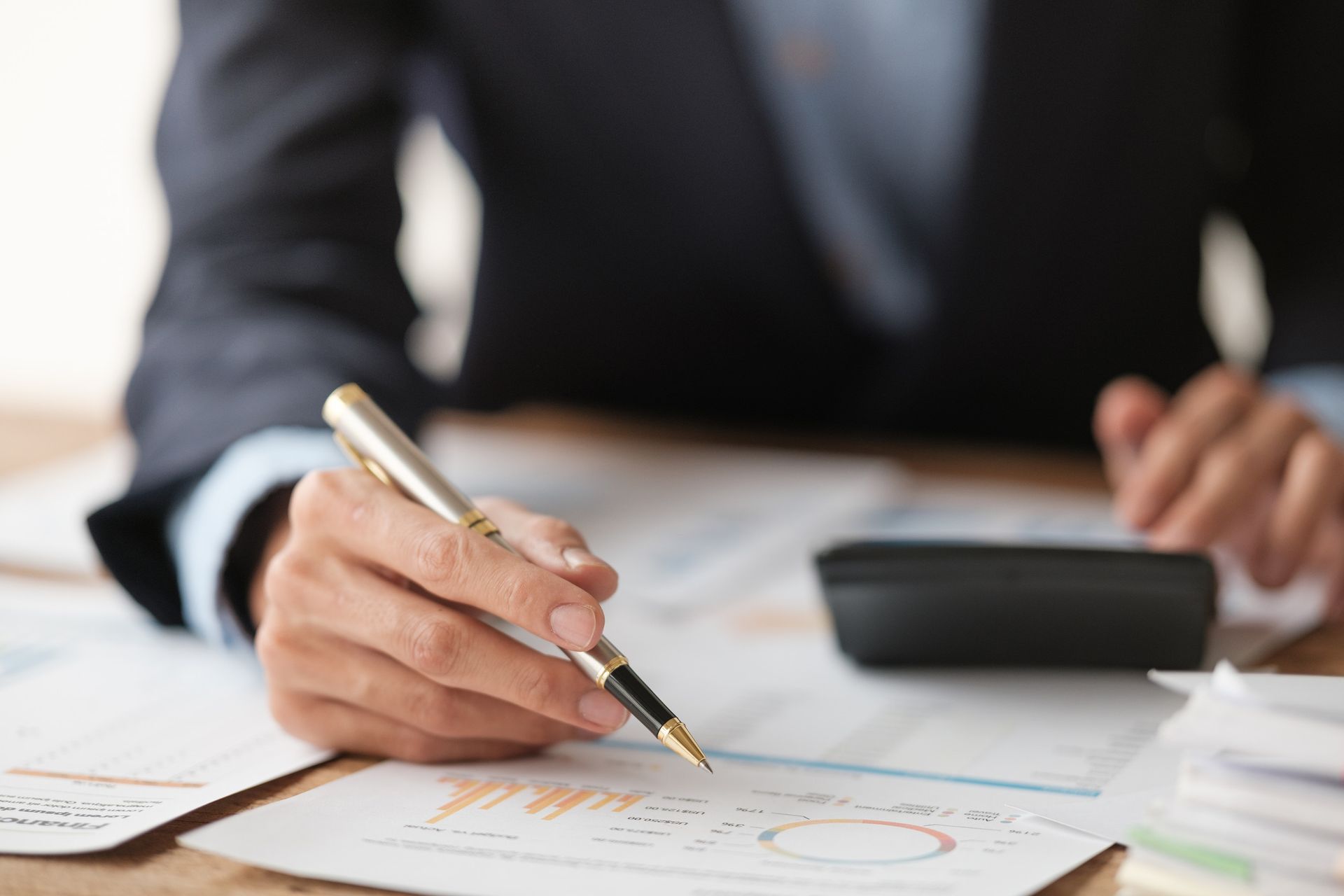 Persona vestida de traje señalando con un bolígrafo unos documentos financieros con una calculadora cerca.